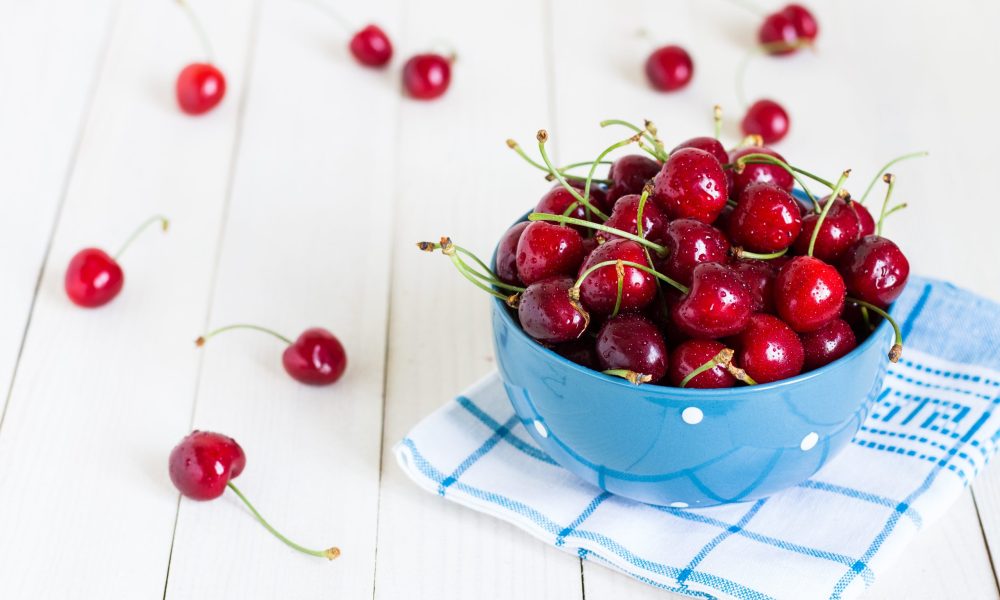 Red cherries in bowl on white wooden background on blue towel. Berry healthy snack
